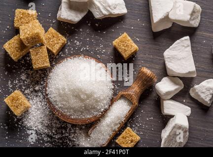 Granulated table sugar in wooden bowl and in the spoon and sugar cubes around it on the table. Stock Photo