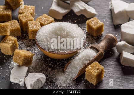 Granulated table sugar in wooden bowl and in the spoon and sugar cubes around it on the table. Stock Photo