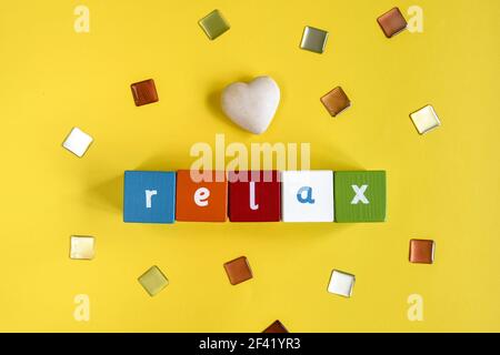 RELAX - Word made from colored wooden blocks. Painted cubes with letters and one stone heart on yellow background. Top view. Copy space. Stock Photo