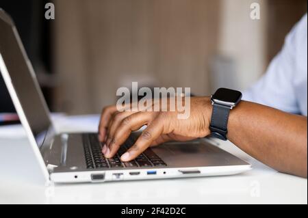 Side profile cropped photo of afro successful businessman hands with fitness watch, typing on keyboard of laptop, side view of the workdesk with modern grey computer, busy typing the data Stock Photo