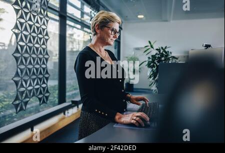 Senior businesswoman working at standing desk. Woman employee working on desktop computer at ergonomic standing desk. Stock Photo