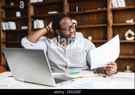 Latin businessman sits at desk in home office near laptop, touches back of the head, thinks over strategy work, generating new fresh ideas, searching solution, trying to figure out the statistics Stock Photo