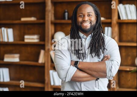 Portrait of a young smiled broad-shouldered african unshaven man with dreadlocks stands with arms on his chest, looks at camera, against the background of shelves with books, successful freelancer Stock Photo