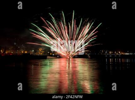 Nautical fireworks. These explode on the surface of water, rather than in the air. City buildings are in the background. Reflection on the water. Stock Photo