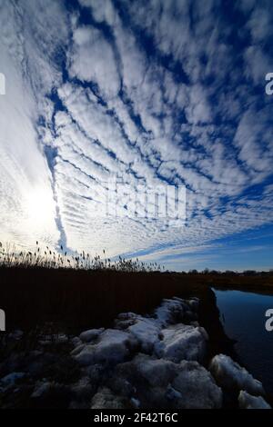 Puffy Cirrus clouds in a graphic arrangement against a bright blue sky along a brook with ice covered rocks and snow. Stock Photo