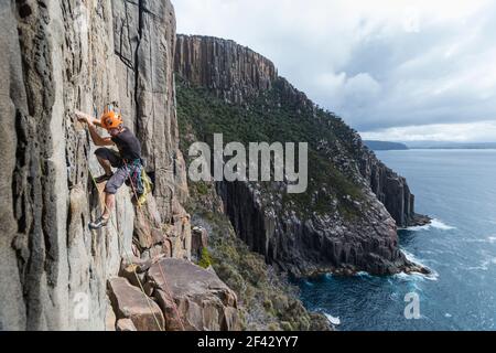 Male rockclimber climbs the exposed edge of a dolerite sea cliff using two ropes as protection in a cloudy day, with bushy cliffs and the ocean in the background in Cape Raoul, Tasmania Stock Photo