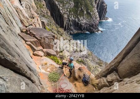 Woman belaying on the exposed edges of a dolerite sea cliff using two ropes as protection in a cloudy day, with bushy cliffs and the ocean in the background in Cape Raoul, Tasmania, Australia Stock Photo