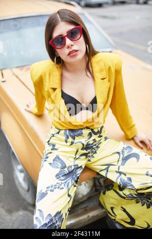 young woman in yellow dress leaning against a car looking at camera Stock Photo