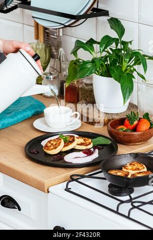 Women's hands pour tea into a cup. cheese pancakes with jam on a Stock Photo