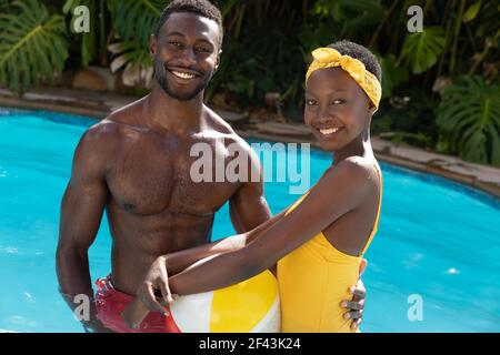 Portrait of african american couple in swimming pool holding beach ball on sunny garden terrace Stock Photo