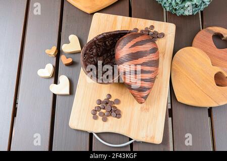Bitter chocolate Easter egg with almond crunch, decorated with edible paint, on a wooden board. Surrounded by chocolate callets and wooden hearts (top Stock Photo