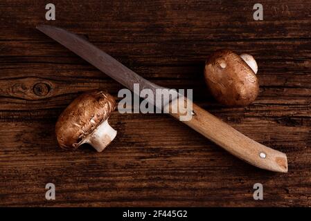 Cremini mushrooms and old kitchen knife on textured wooden table. Two baby portobello brown mushrooms. Closeup. Minimal style Stock Photo