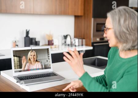Senior mature middle-aged woman in glasses sitting in the kitchen, waving at laptop, chatting looking at the screen with female friend image, staying connected in pandemic, self-isolation concept Stock Photo