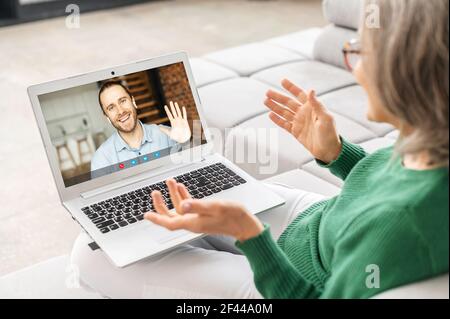 Close-up of laptop and senior mature woman grandmother in glasses sitting in the kitchen, talking and looking at screen with male friend son image, gesturing, staying at home, connected in pandemic Stock Photo