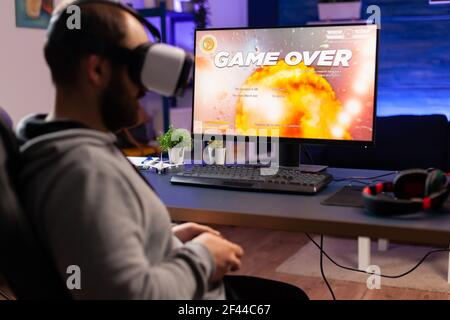 Over shoulder of focused cyber player wearing virtual reality headset during online championship. Game over for pro gamer using console for shooter gaming competition late at night in living room Stock Photo