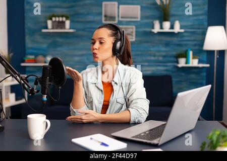 Vlogger giving flying kiss while making daily online video blog. Digital influcencer recording talk show at home studio brodcast using headphones, professional podcast microphone and modern laptop Stock Photo