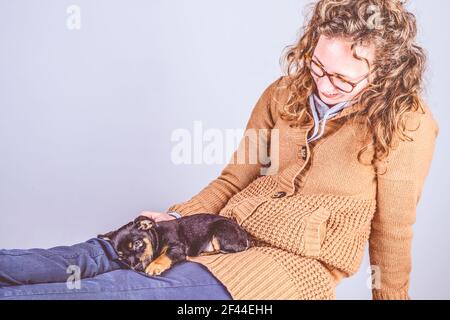 Detail of a beautiful woman with glasses and curly brown hair, Sitting smiling, with a nearly sleeping Jack Russel Terrier puppy on her legs. In Stock Photo