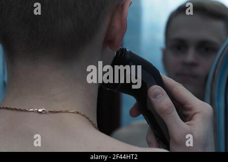 A young man shaves his face with a razor and looks in the mirror. Stock Photo