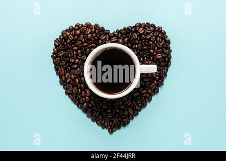 Coffee beans in shape of heart with white cup on blue background. Creative minimal coffee drink concept. Stock Photo