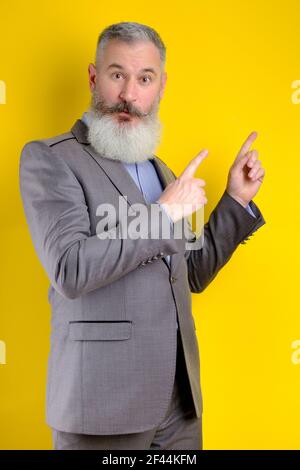 Studio portrait mature businessman dressed in gray suit points aside, I choose you this, yellow backdrop Stock Photo