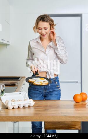 Happy young woman holds skillet with fried eggs and toasts in hand Stock Photo