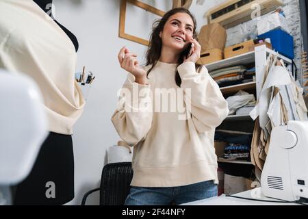 Smiling nice brunette girl seamstress talking on mobile phone while working in atelier Stock Photo