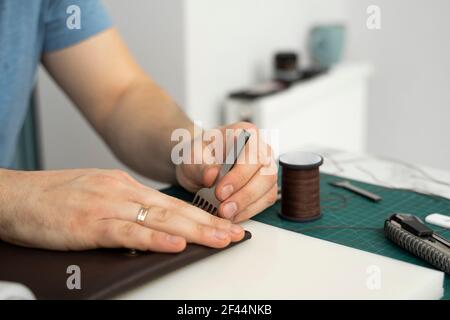 Mens hand holding a leather carving hammer and steel chisel hole punch and makes a leather wallet in his workshop. Working process with a brown Stock Photo