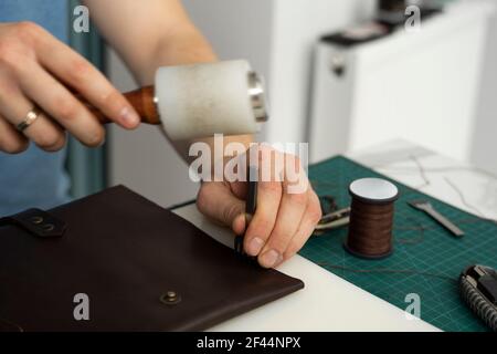 Mens hand holding a leather carving hammer and steel chisel hole punch and makes a leather wallet in his workshop. Working process with a brown Stock Photo