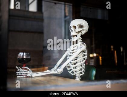 Human skeleton bones sat in leather chair in pub holding a glass of alcohol wine waiting a long time for pubs and bars to re-open after being closed Stock Photo