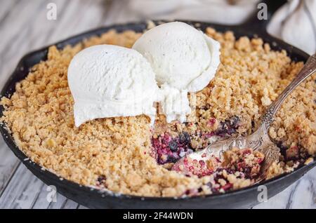 Blackberry and Blueberry Cobbler baked in a cast iron pan and topped with a golden oatmeal crisp with vanilla ice cream over a rustic white wood table Stock Photo
