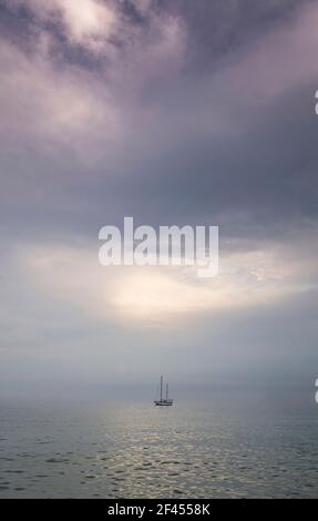 a small sailboat anchored in the middle of a sea in total calm in a sunset with a cloudy sky in mauve tones, feeling of calm, isolation, minimalism an Stock Photo