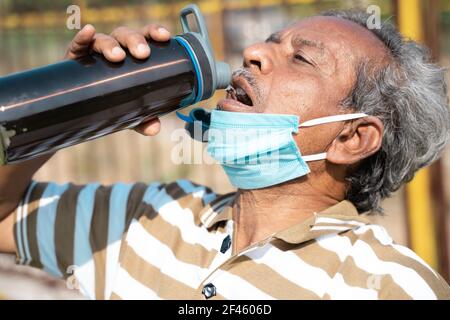 old man drinking water from bottle by placing mask below face during hot sunny day - Concept of healthcare, medical, thirsty on outdoors during Stock Photo