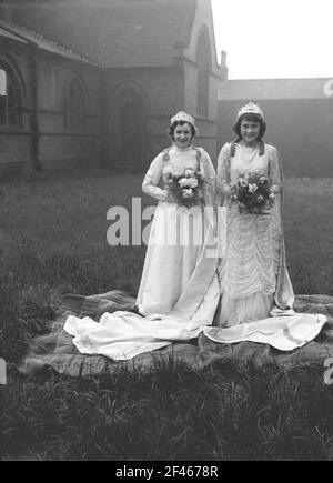 1956, historical, two teenage girls standing outside in the grounds of a church each wearing long, elaborate lace gowns and holding bouquets of flowers before taking part in the May Day parade, a popular day of celebration in much of northern England in tihs era. On their heads are crowns, with the girl on the right having been chosen as this year's May Queen. A popular day of celebration marking the beginning of summer, May first, commonly known as May Day, is an ancient tradition dating back to the celebration of the roman goddess of flowers, Flora Stock Photo