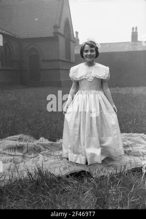1956, historical, a young teenage girl standing on a rug outside in the grounds of a church wearing a long, elaborate lace gown, before taking part in the May Day parade, a popular day of celebration marking the beginning of summer. May first, commonly known as May Day, is an ancient tradition dating back to the celebration of the roman goddess of flowers, Flora. Stock Photo