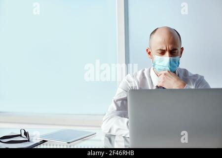 thoughtful businessman in a protective mask sitting in front of an open laptop. Stock Photo