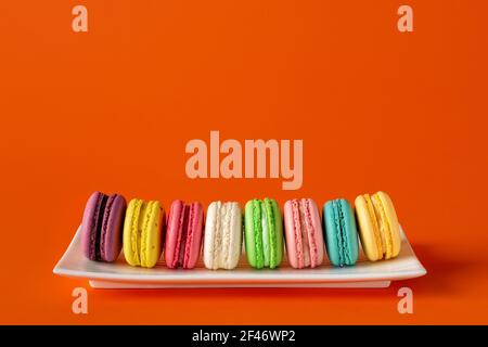 Multicolored macaroons in a row on a white rectangular plate against bright red background. Mockup of greeting card with colorful macaron cakes. Stock Photo