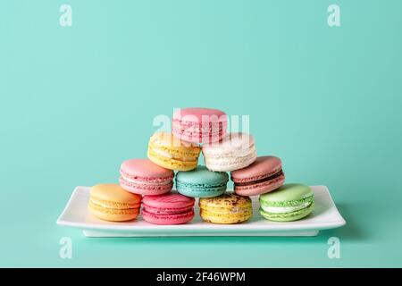 Colorful macaroons pyramid on a white rectangular plate against pastel turquoise background. Mockup of greeting card with sweet multicolored macarons. Stock Photo