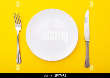 Ceramic white plate with knife and fork on yellow surface, kitchen table, lay out, top view, space for text Stock Photo