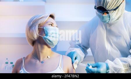 Nurse in protective suit injecting vaccine to young caucasian woman. Covid prevention. High quality photo Stock Photo