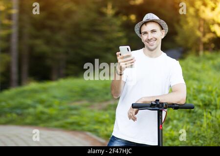 Young man in hat use mobile phone, book online rental electric scooter in city. Innovative transport Stock Photo