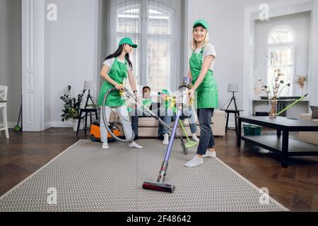 Two female janitors in uniform using modern vacuum for cleaning room. Multiracial men sitting on couch and using tablet on background. Addiction from gadgets. Stock Photo