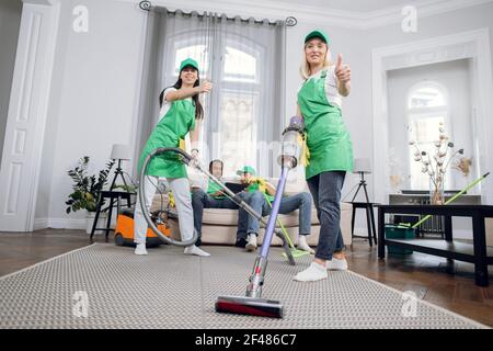 Two female janitors in uniform using modern vacuum for cleaning room. Their colleagues, multiracial men sitting on couch and using tablet on background. Addiction from gadgets. Stock Photo