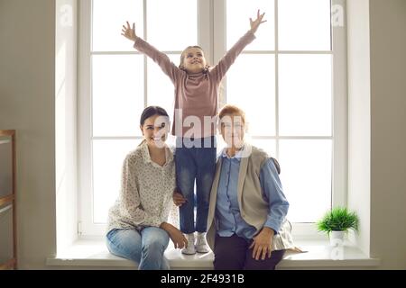 Older woman with her adult daughter and little granddaughter posing while sitting on the windowsill. Stock Photo