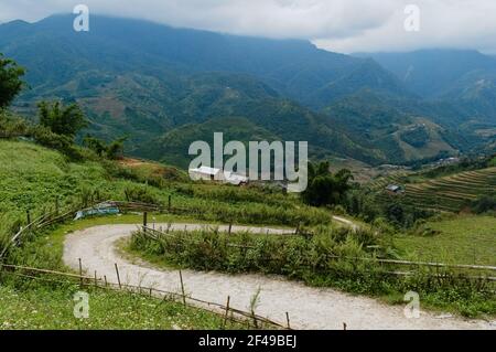 Winding curvy road going down to valley. Rural trail with curves along rice terraces and farm fields. Clouds over mountain range on background. Sa Pa Stock Photo