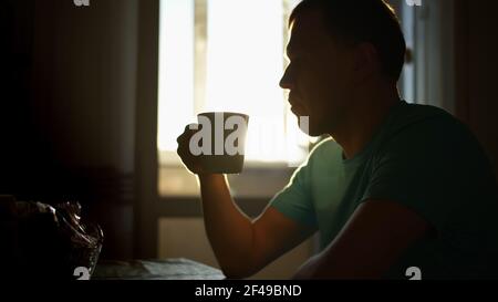 Silhouette of a man in the sunbeams of dawn drinks hot coffee while sitting at home, camera movement