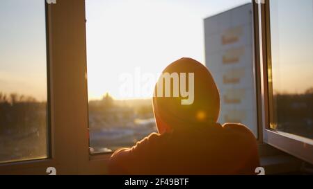 A guy in a hoodie with a hood on his head looks at the sunset while standing in an open window Stock Photo
