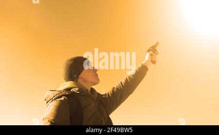 Young man in a jacket and a hat shoots up with a pistol against the background of an orange sky with a white sun Stock Photo