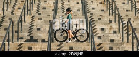 Website header of Young professional female cyclist in cycling garment and protective gear standing on the steps with her bike after having a training Stock Photo