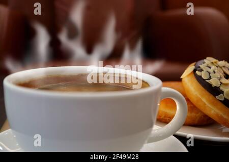 Cup of Hot Coffee with Rising Steam and a Plate of Donuts in Background Stock Photo