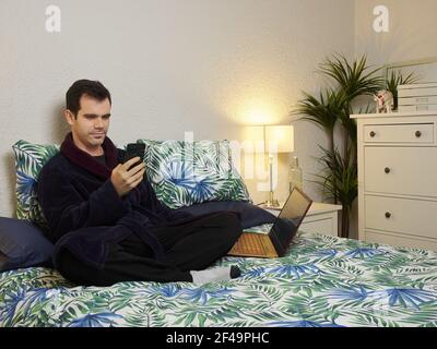 A handsome Hispanic male in a dressing gown sitting on a bed and looking at his phone Stock Photo
